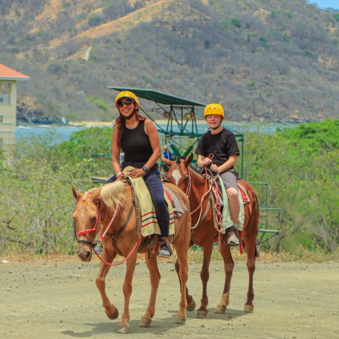 Two people on horseback wearing helmets, mountains and a building in the background.