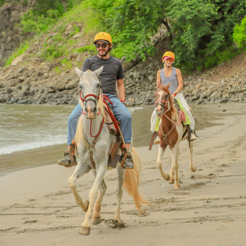 Two people riding horses on a sandy beach with rocky cliffs.