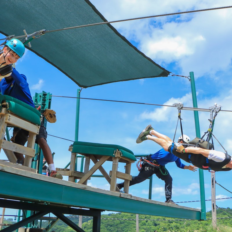 Two people ziplining from a platform in harnesses, wearing helmets, under a blue sky.