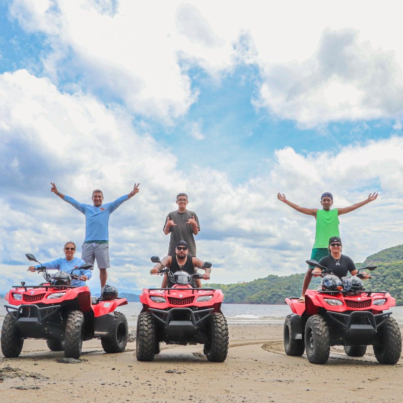 Four people on red ATVs on a beach, two standing, with arms outstretched; hills and cloudy sky in background.