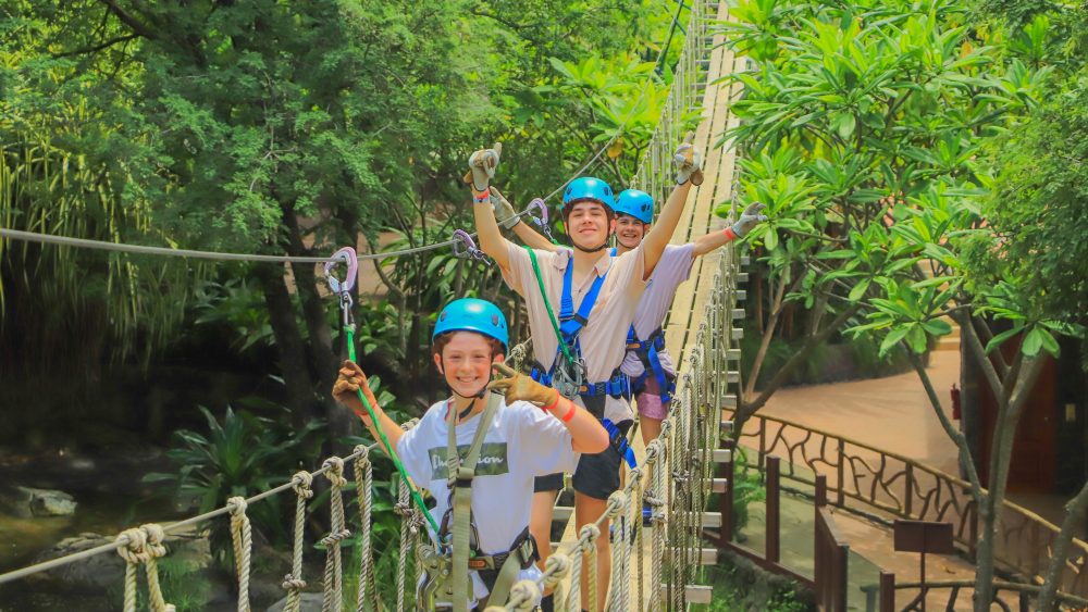 Three people in blue helmets crossing a rope bridge in a lush, green forest.