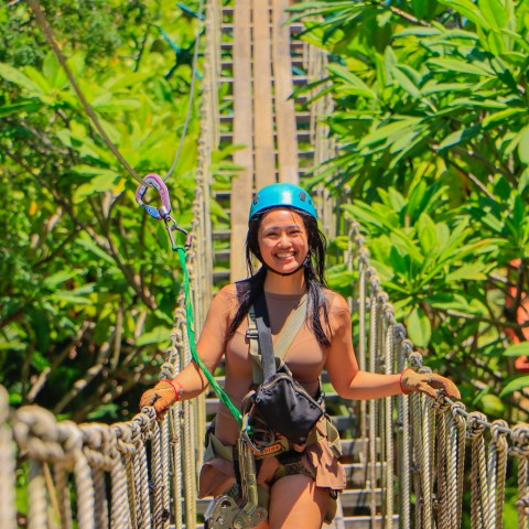 Person with helmet and gloves on a rope bridge surrounded by lush greenery.