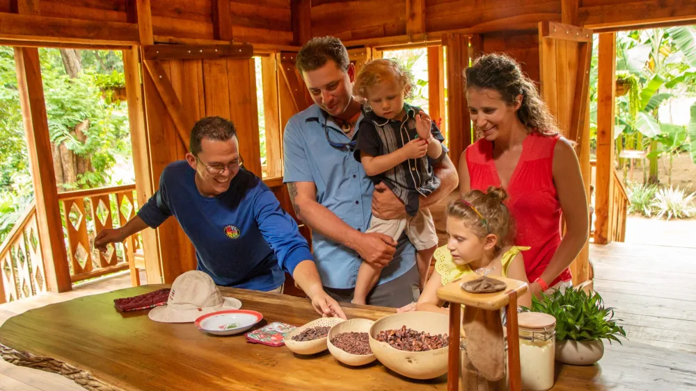 Family exploring cocoa beans with a guide at a wooden table in a rustic setting. --- Children learning to make chocolate during Diamante’s Cultural Experience