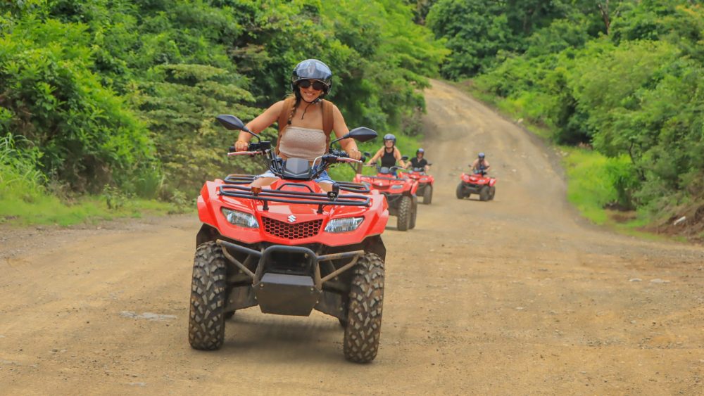 People on red ATVs riding on a dirt road through lush green forest.