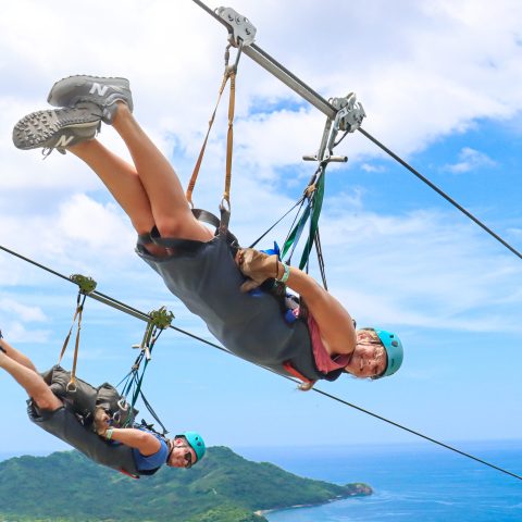 Two people zip-lining over a scenic landscape with ocean and hills in the background.