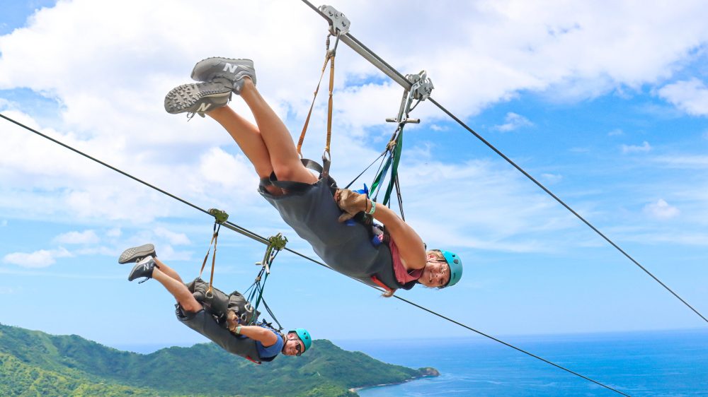 Two people zip-lining over a scenic landscape with ocean and hills in the background.