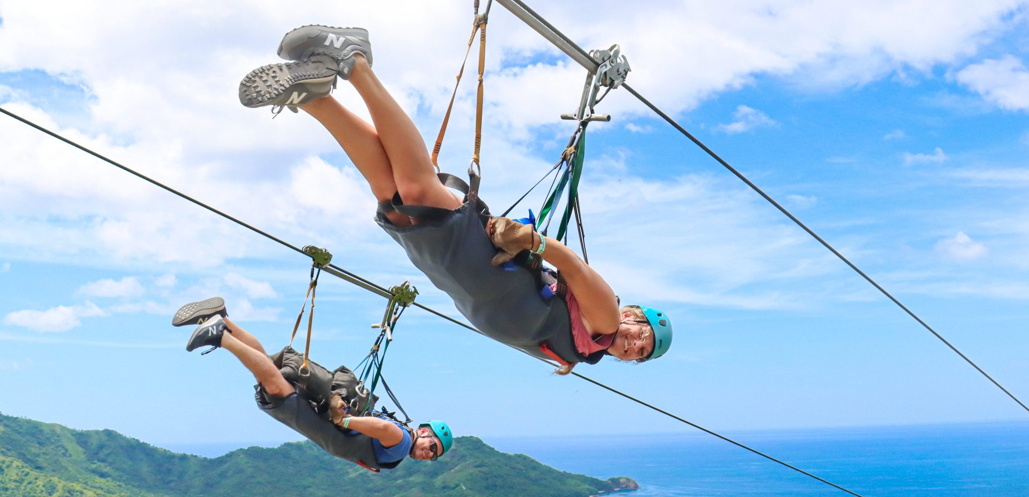 Two people zip-lining over a scenic landscape with ocean and hills in the background.