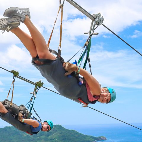 Two people zip-lining over a scenic landscape with ocean and hills in the background.