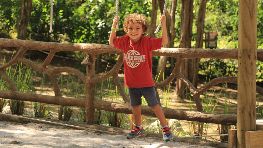 Child in a red shirt playing on rope swings at a park with wooden fence and greenery.