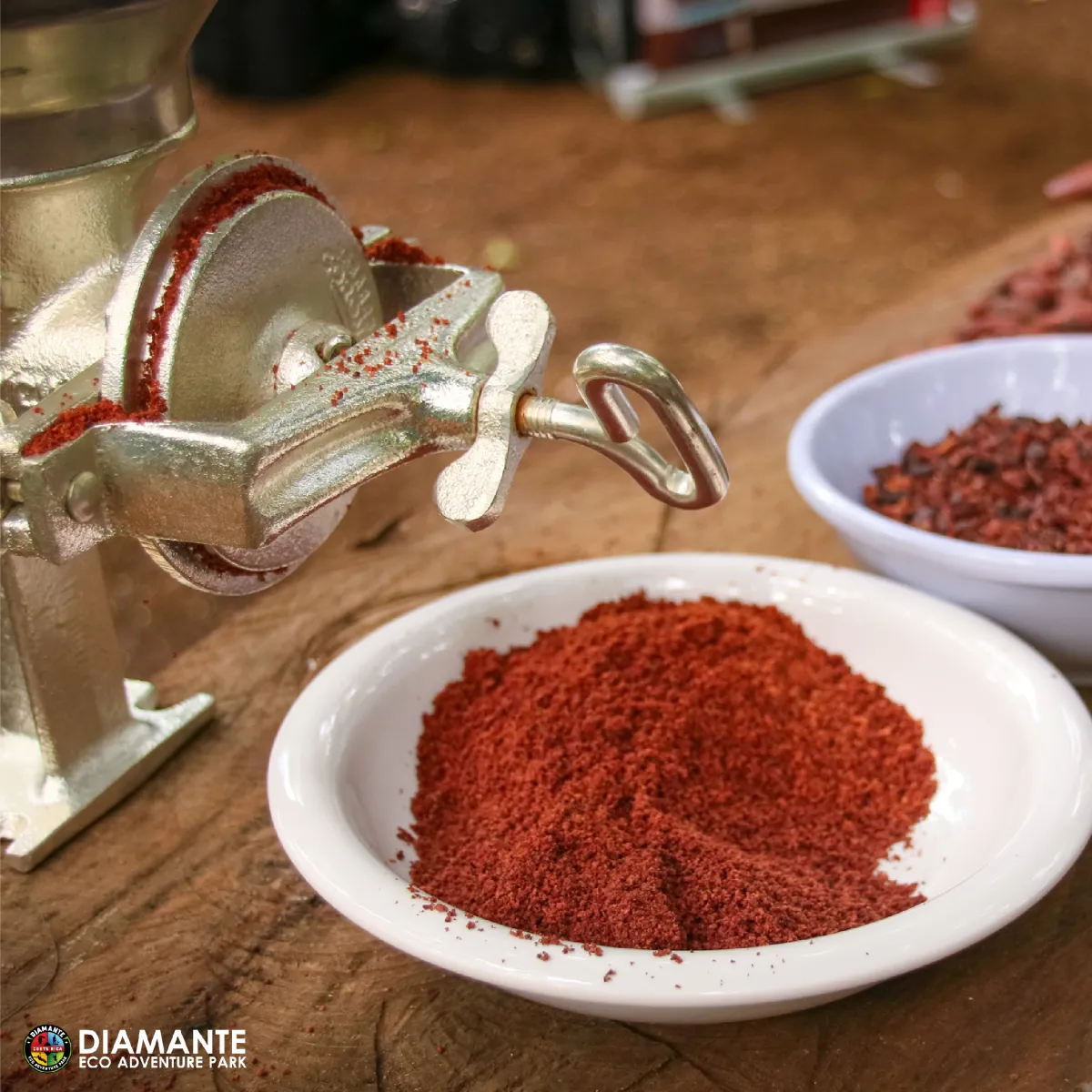Silver grinder with red spice powder in white dish on wooden surface.