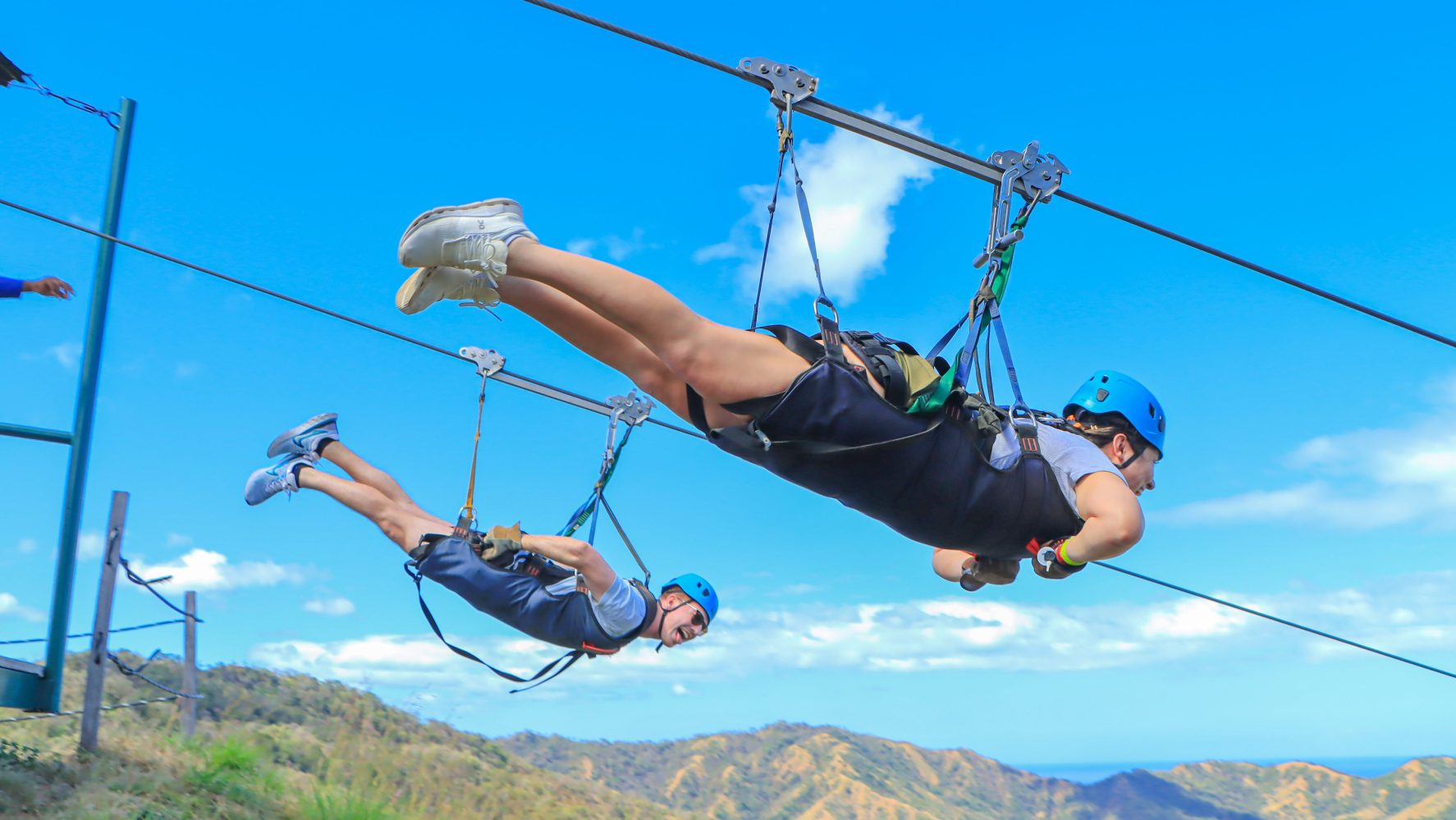 Two people with helmets riding a zip line over a scenic mountain landscape under a clear blue sky.