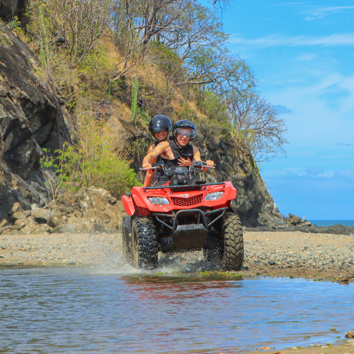 Two people riding a red ATV through shallow water near rocky cliffs and the ocean.