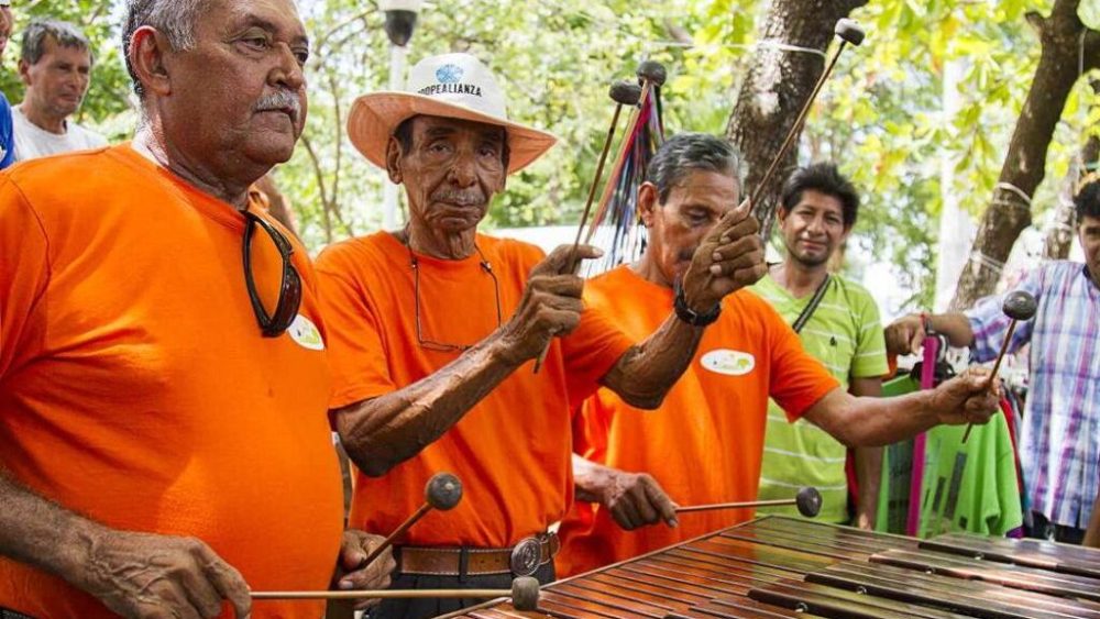 Three men in orange shirts playing a marimba outdoors, surrounded by trees and onlookers.