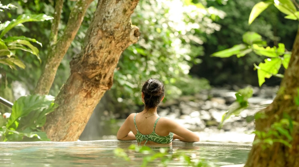 Person in a green swimsuit relaxing in a natural hot spring, surrounded by lush greenery.
