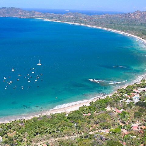 Aerial view of a coastal town with boats in a blue ocean bay and green hills in the background.