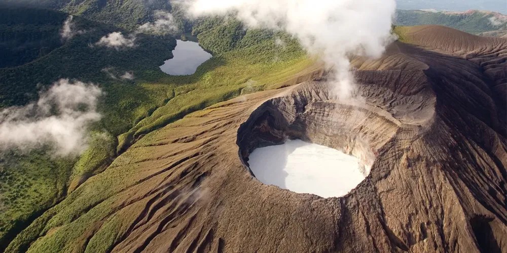 Aerial view of a volcano crater with steam, surrounded by lush greenery and a lake nearby.
