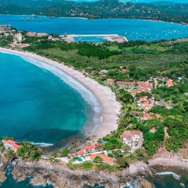 Aerial view of a beach with turquoise waters, surrounded by green hills and buildings.