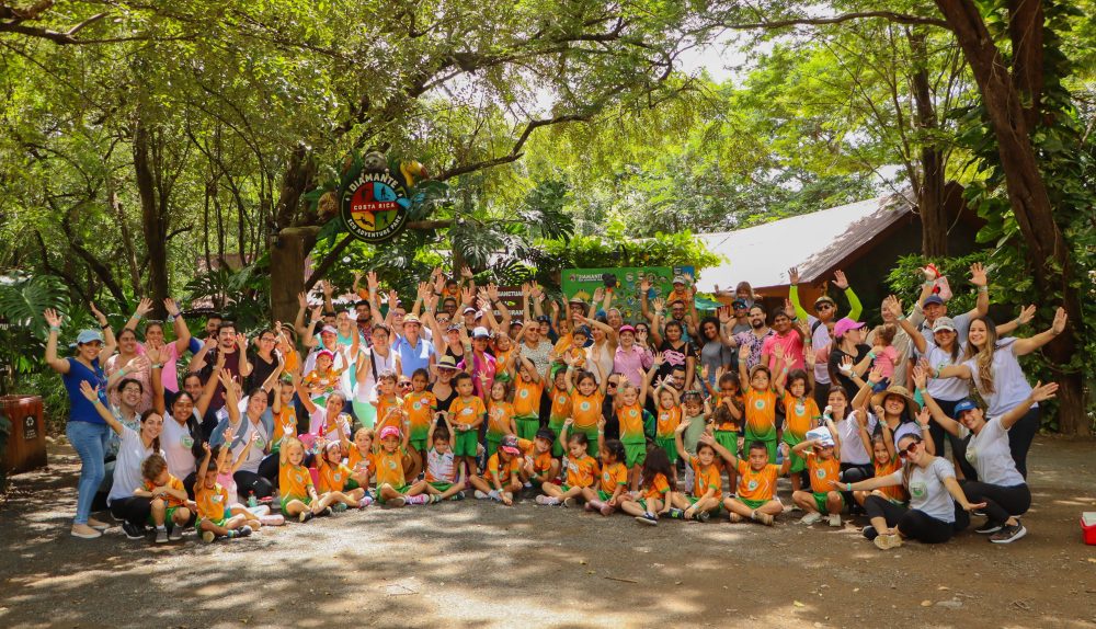 Large group of children and adults posing outdoors under trees, smiling and waving.
