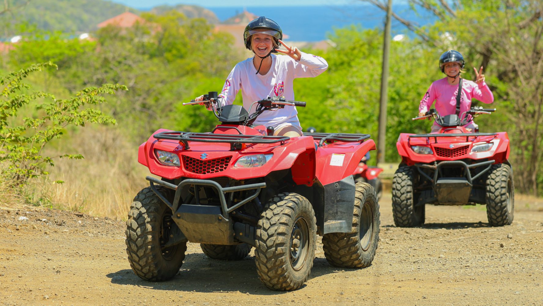 Two people riding red ATVs on a dirt path with greenery and ocean in background.