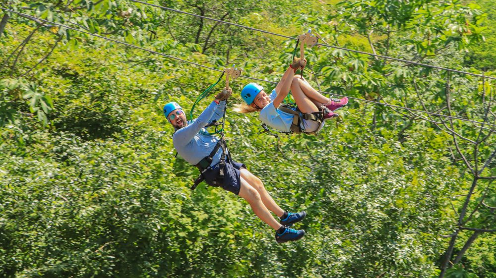 Two people zip-lining through a lush green forest, wearing helmets and harnesses.