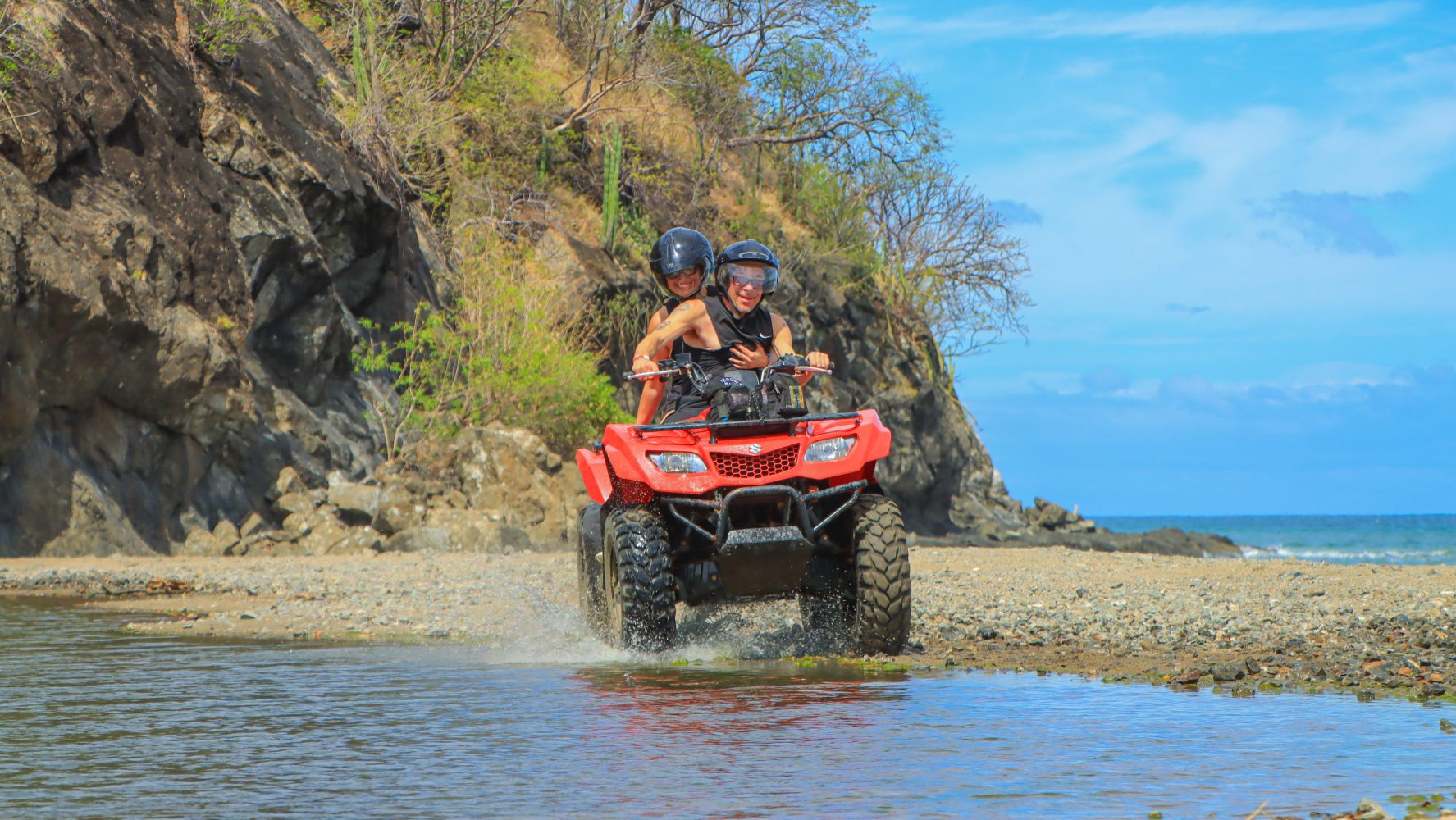 Two people riding a red ATV through water near a rocky coast.